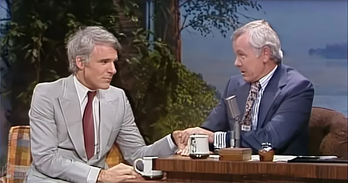 Two men in suits are seated at a table, engaged in conversation on a talk show set.