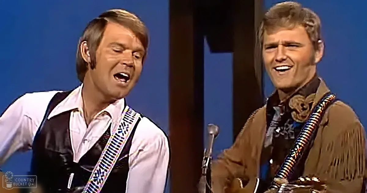 Two men in Western attire sing and play guitars on stage, one in a white shirt and black vest, the other in a brown...