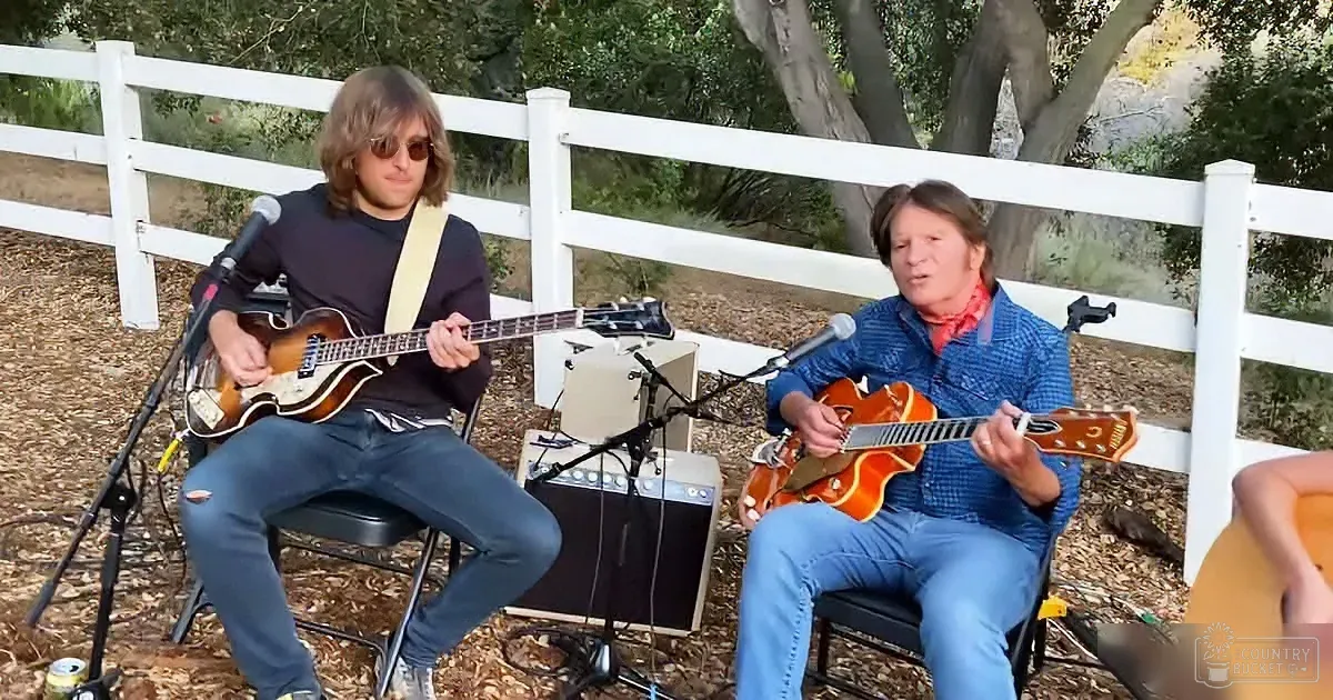 Two musicians play guitars outdoors, one with a bass, surrounded by microphones and amplifiers.