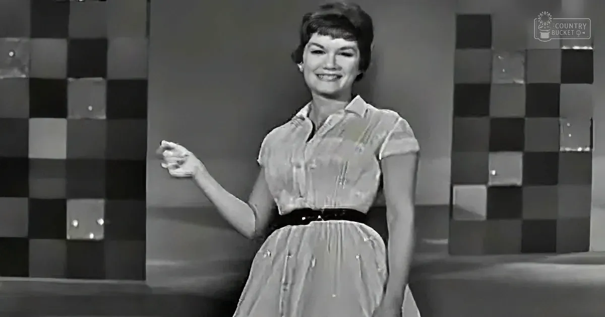A woman in a 1950s-style dress smiles, gesturing with her right hand, against a checkered backdrop.