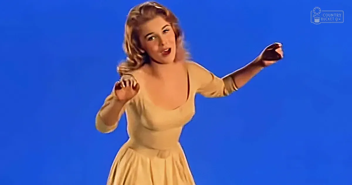 A woman in a beige dress sings with raised arms against a blue background.