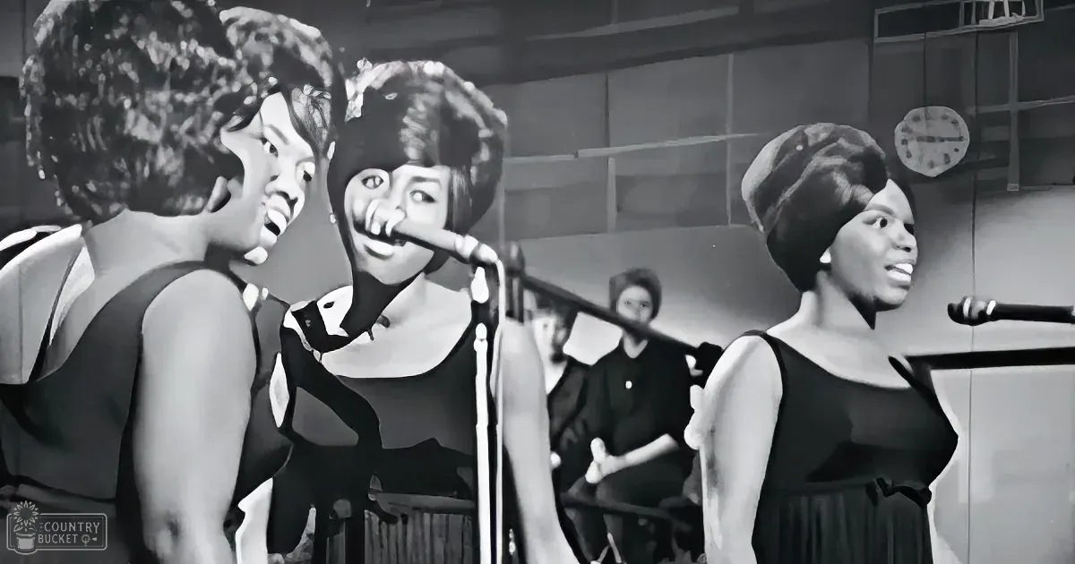 A woman in a black dress sings into a microphone, accompanied by two other women in similar attire.