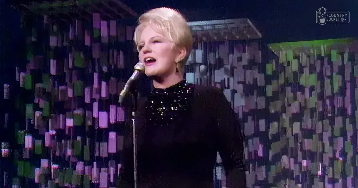 A woman in a black sequined dress sings into a microphone on stage with a colorful geometric backdrop.