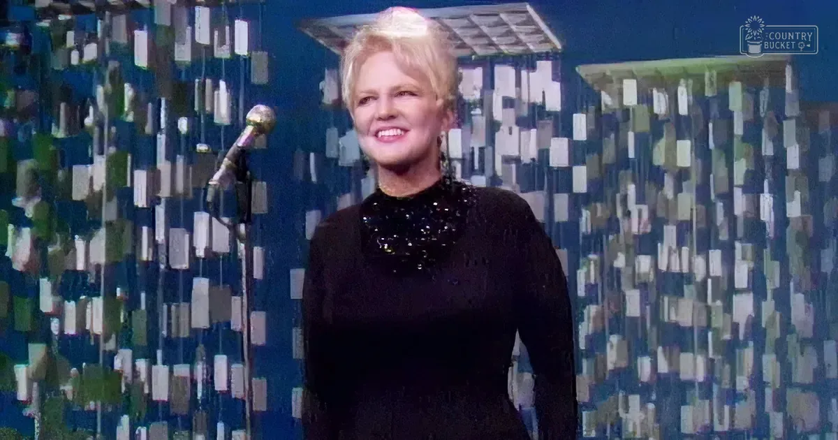 A woman in a black sequined dress smiles on stage with a microphone and patterned backdrop.