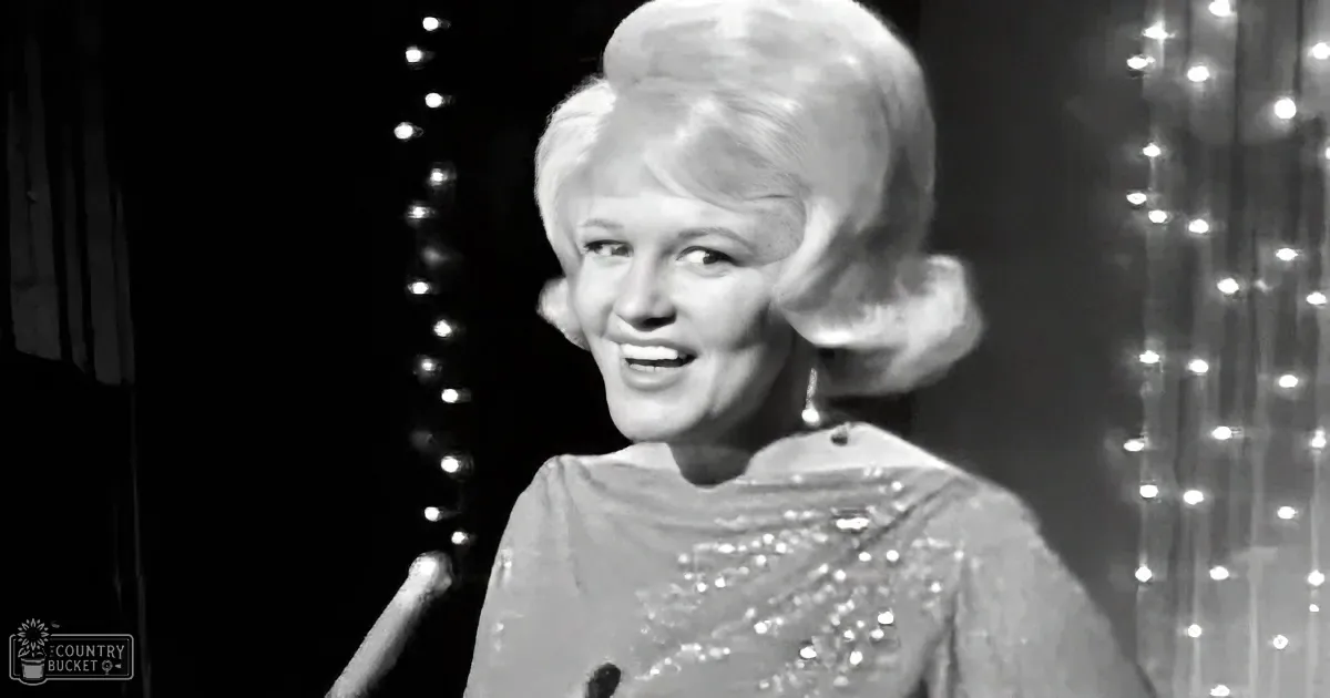 A woman with blonde hair, wearing a sparkly dress, smiles while holding a microphone against a backdrop of twinkling lights.
