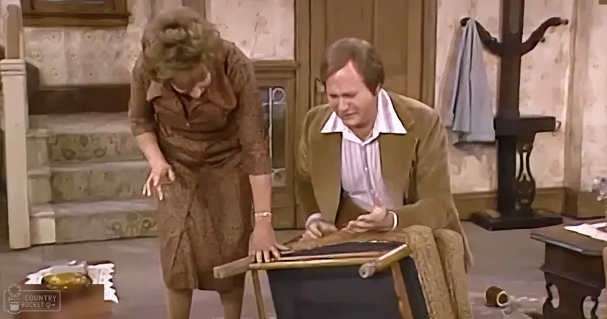 A woman in a brown dress bends over, while a man in a brown jacket leans on a table, both in a room with stairs and a...