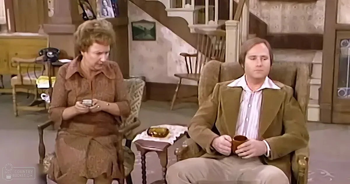 A woman in brown sits in a chair, holding a small object, while a man in a brown suit sits across from her, holding a mug.