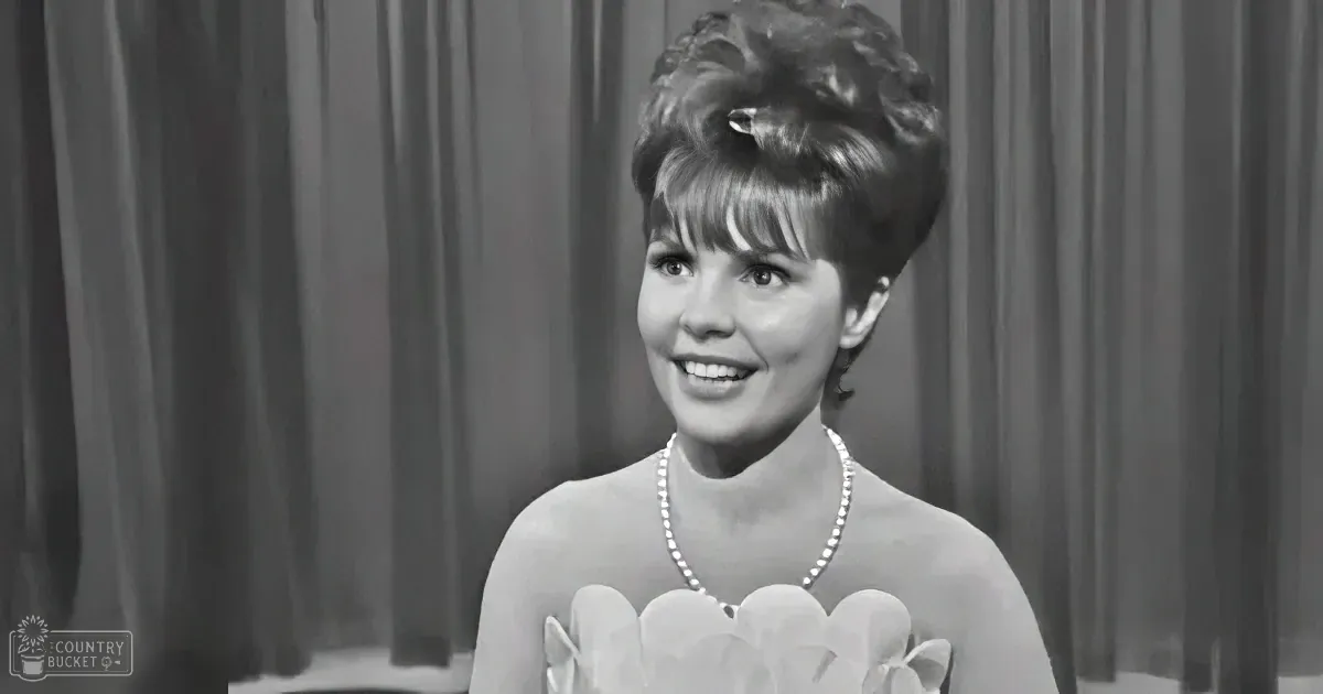 A woman in a floral dress and pearl necklace smiles against a curtain backdrop.