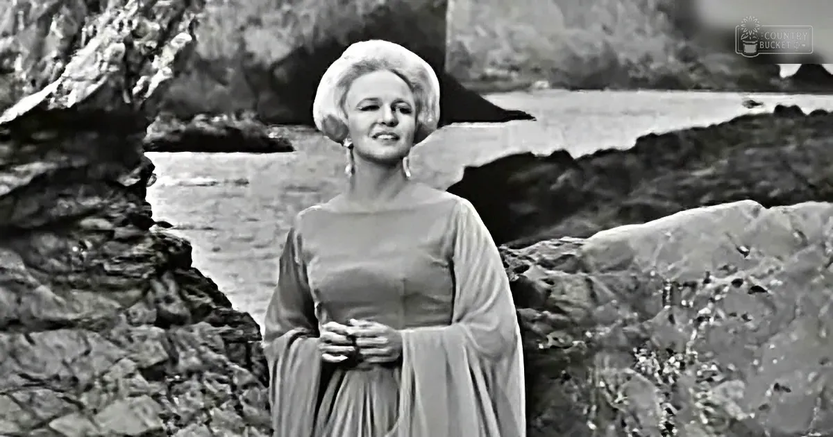 A woman in a flowing dress and large hat stands amidst rocky terrain, smiling.