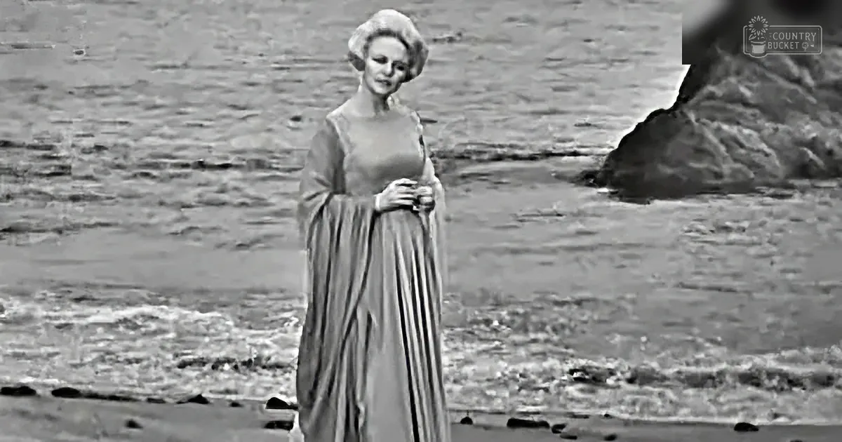 A woman in a flowing dress stands on a beach, holding her skirt, with a rocky outcrop in the background.
