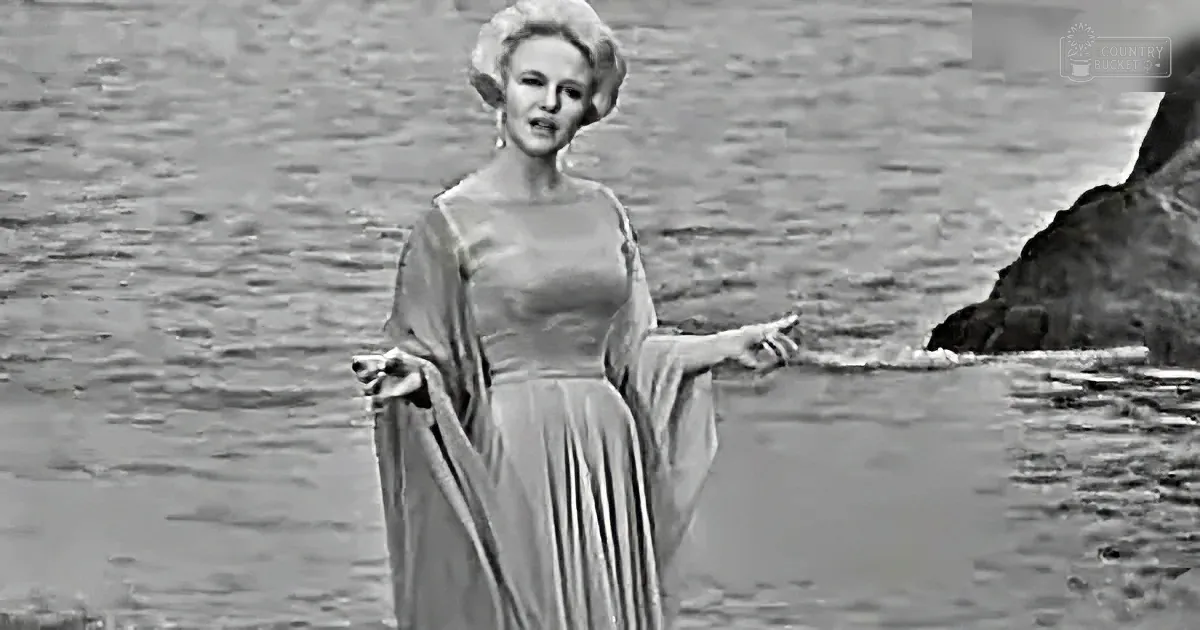 A woman in a flowing dress stands by water, arms outstretched, in a dramatic pose.