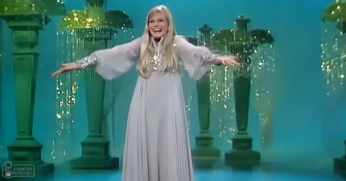 A woman in a flowing white gown with arms outstretched stands on a stage with green pillars and sparkling water effects.