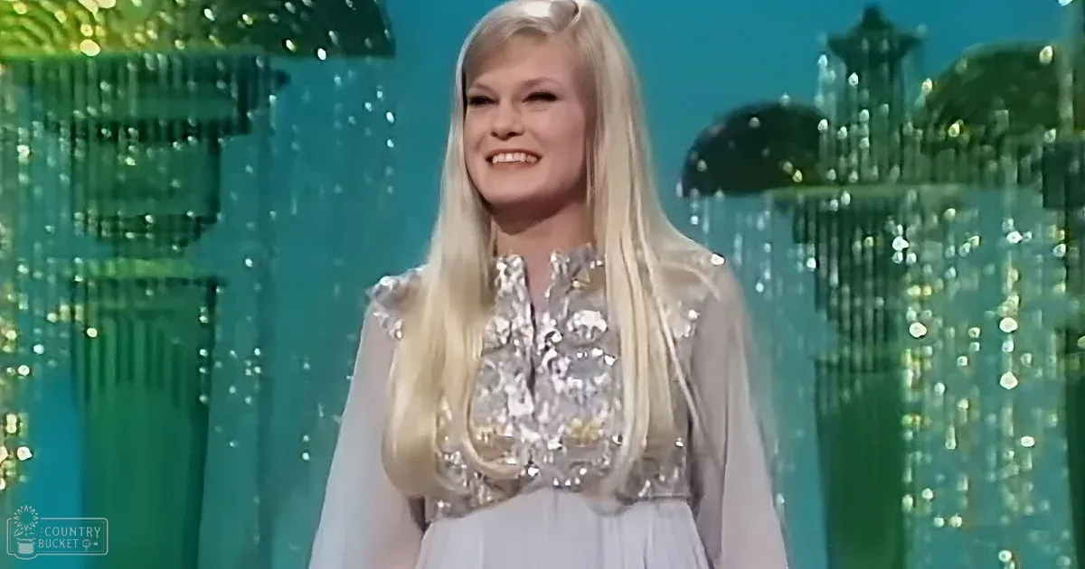 A woman with long blonde hair smiles in a white dress with sequins, standing in front of a glittery green background.