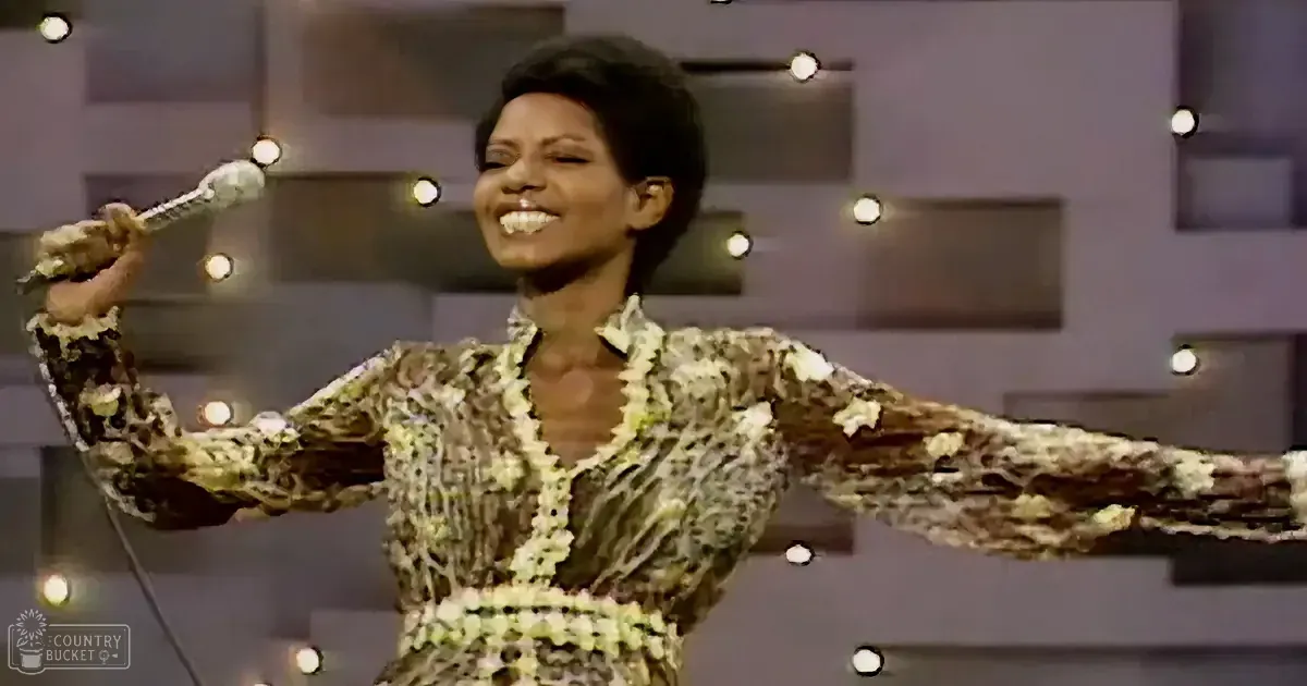 A woman in a patterned dress holds a microphone, smiling, with outstretched arms, against a backdrop of lights and a dark...