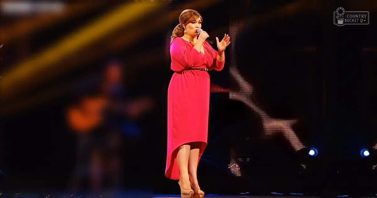 An older adult woman speaks into a microphone in a dimly lit stage setting with an audience clapping. She wears a black and white floral-patterned top.
