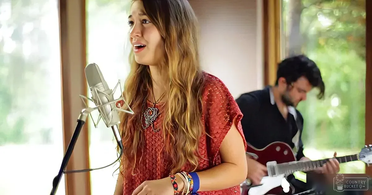 A woman in a red dress sings into a microphone, accompanied by a guitarist in the background.