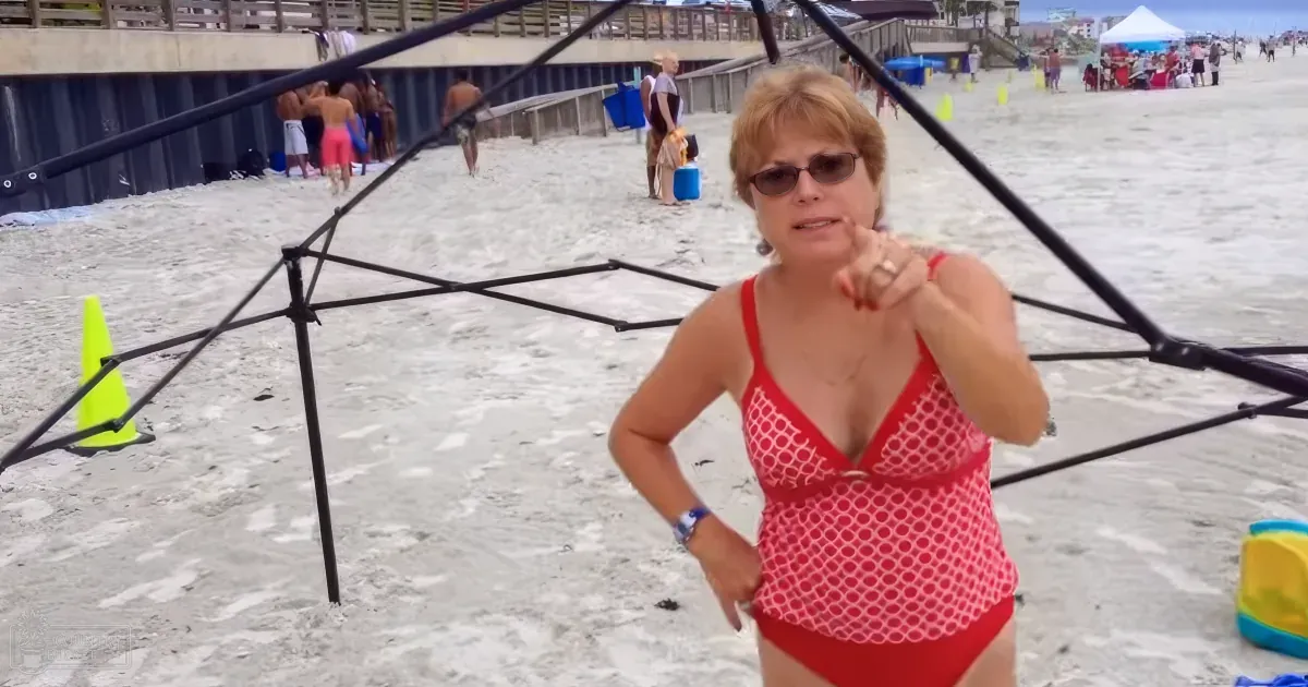 A woman in a red swimsuit stands on a beach, pointing forward, with a black umbrella and yellow cone nearby.