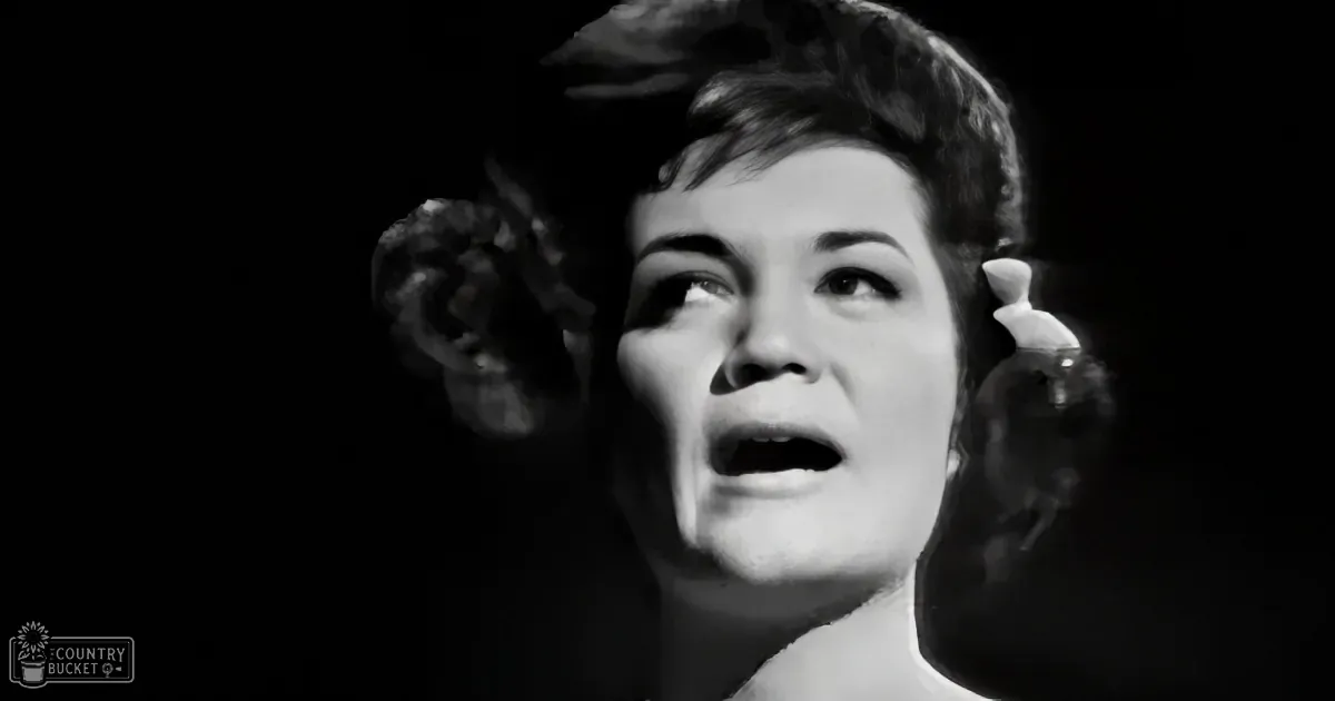 A woman with short hair and floral earrings is singing, her mouth open, against a dark background.