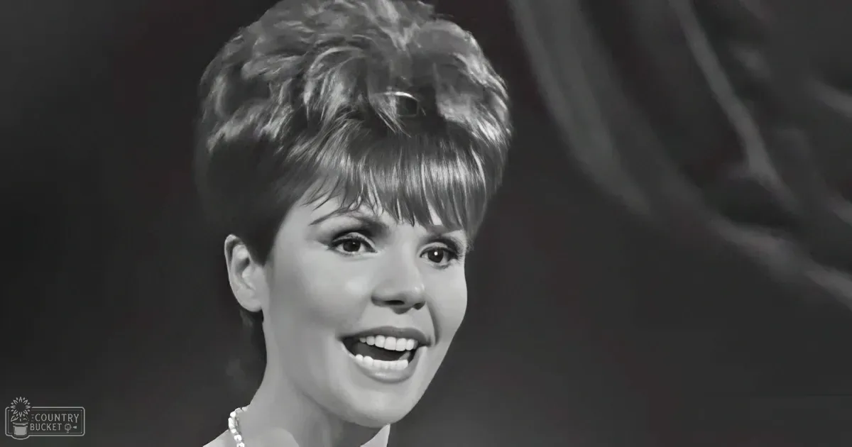 A woman with short, styled hair and a pearl necklace smiles warmly, set against a dark background.