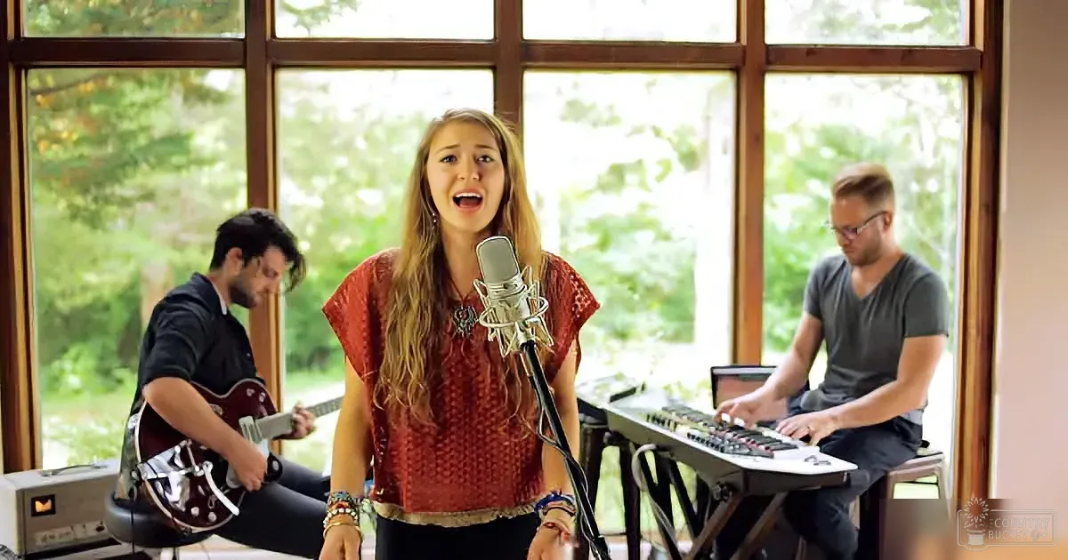 A woman sings into a microphone, accompanied by a guitarist and keyboardist in a room with large windows.