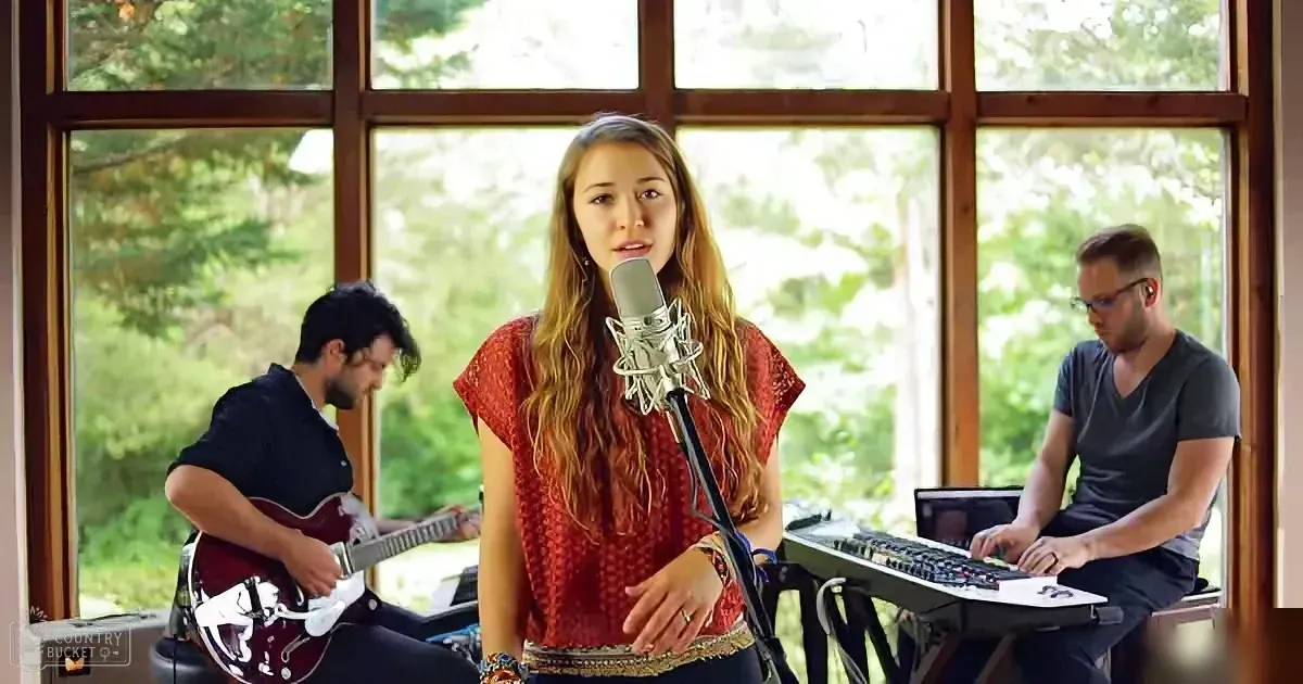 A woman sings into a microphone, accompanied by a guitarist and keyboardist in a room with large windows.