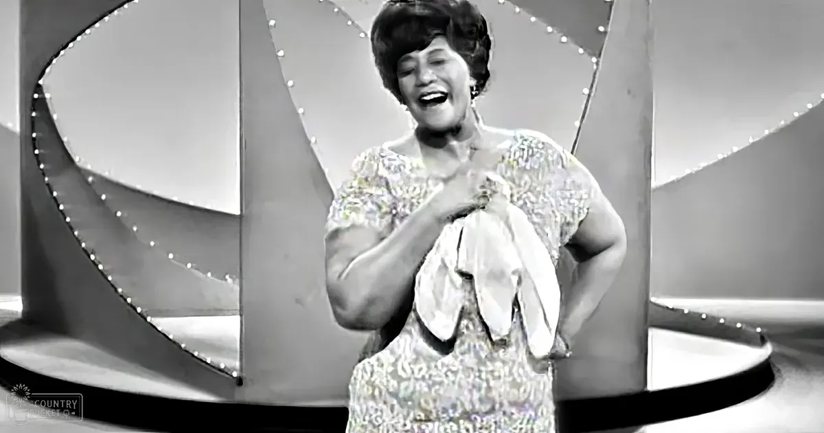 A woman in a sparkling white dress smiles and gestures with her hands on a stage with a decorative backdrop.
