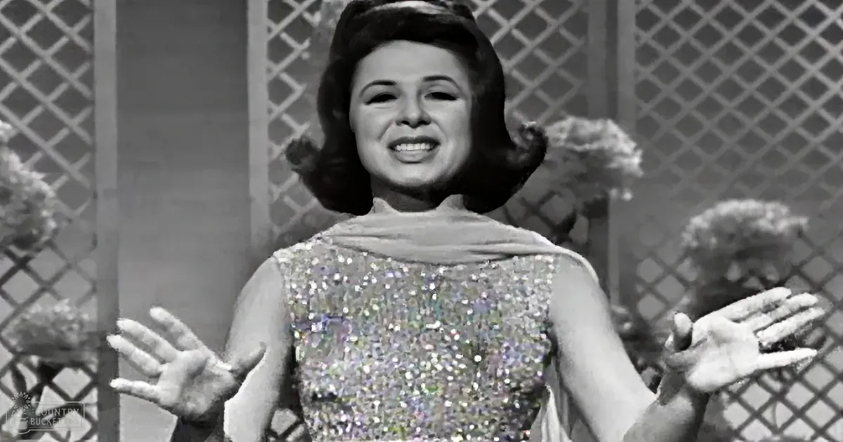 A woman in a sparkly dress sings with her hands raised, surrounded by lattice and flowers.