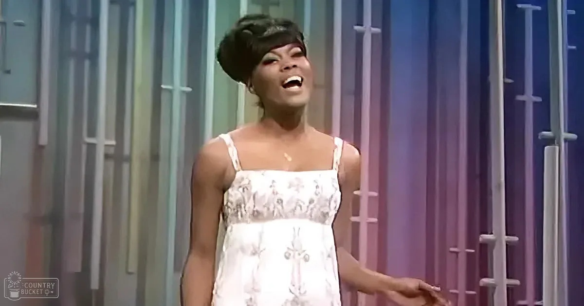 A woman in a white dress with a floral pattern is singing on stage with a colorful background.