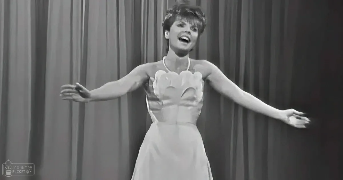 A woman in a white dress with a heart-shaped neckline is singing with arms outstretched.