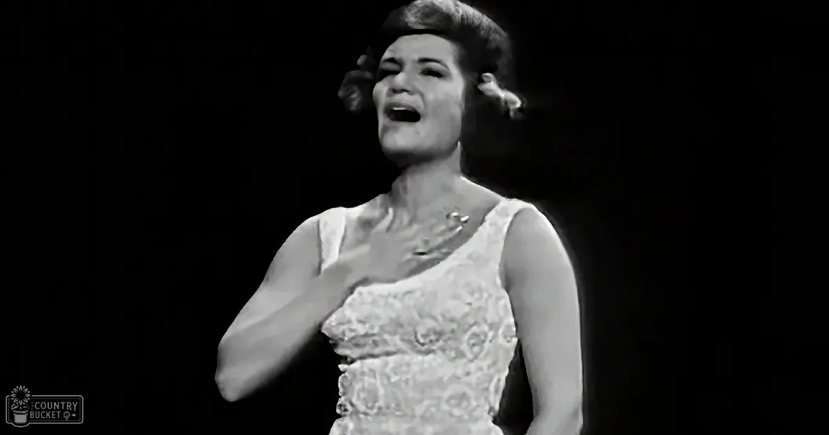 A woman in a white dress is singing passionately with her hand on her chest.