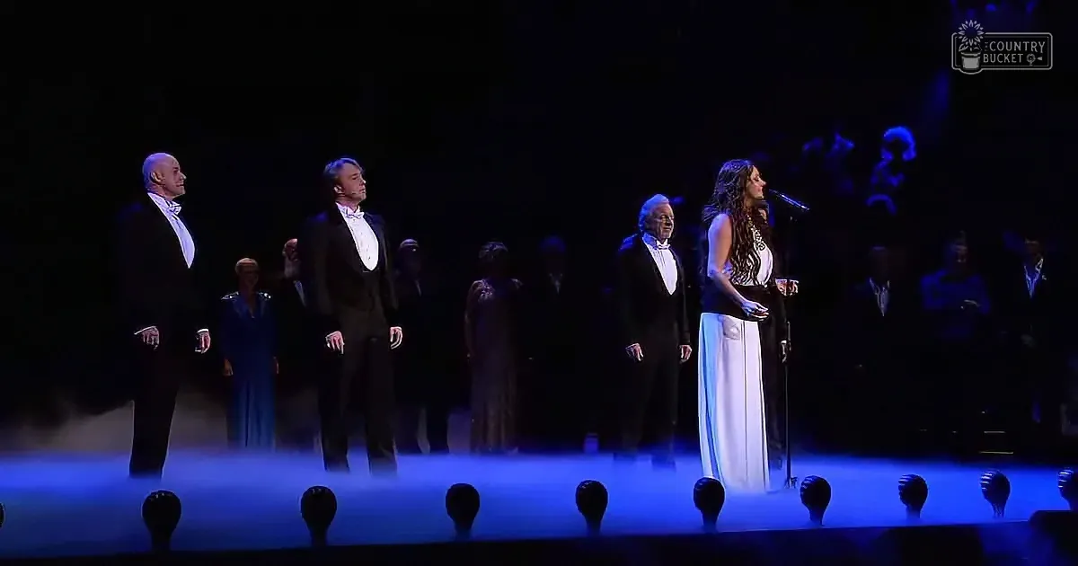 A woman in a white dress sings on stage with a man in a tuxedo, surrounded by other formally dressed performers.