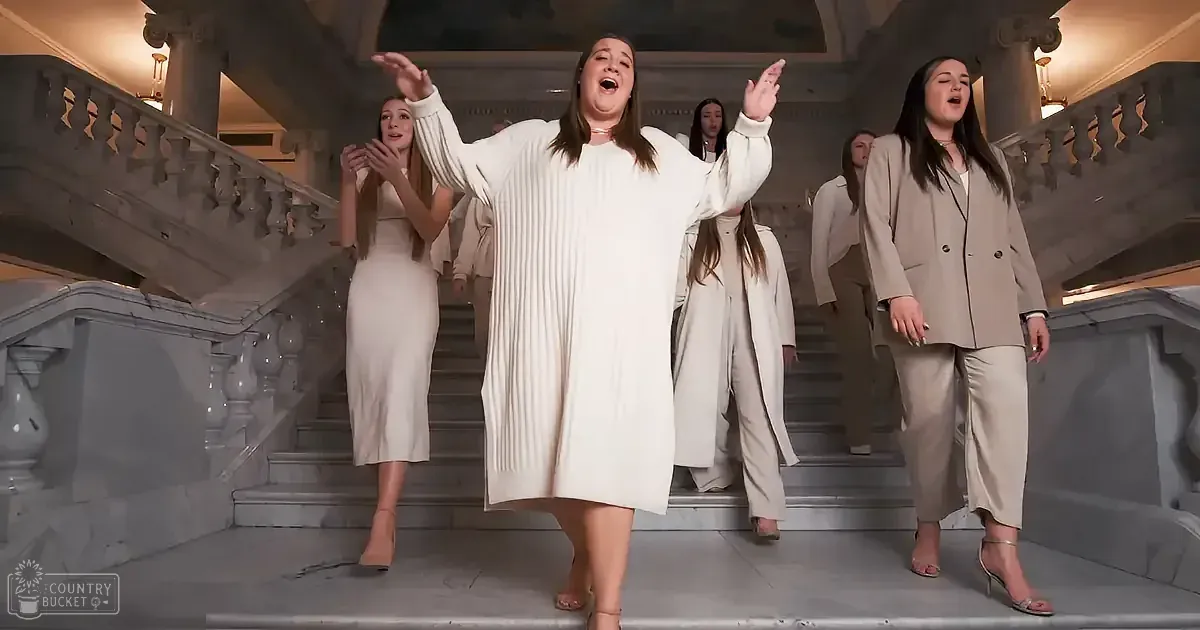 A woman in a white dress stands on a grand staircase, arms raised, singing passionately.
