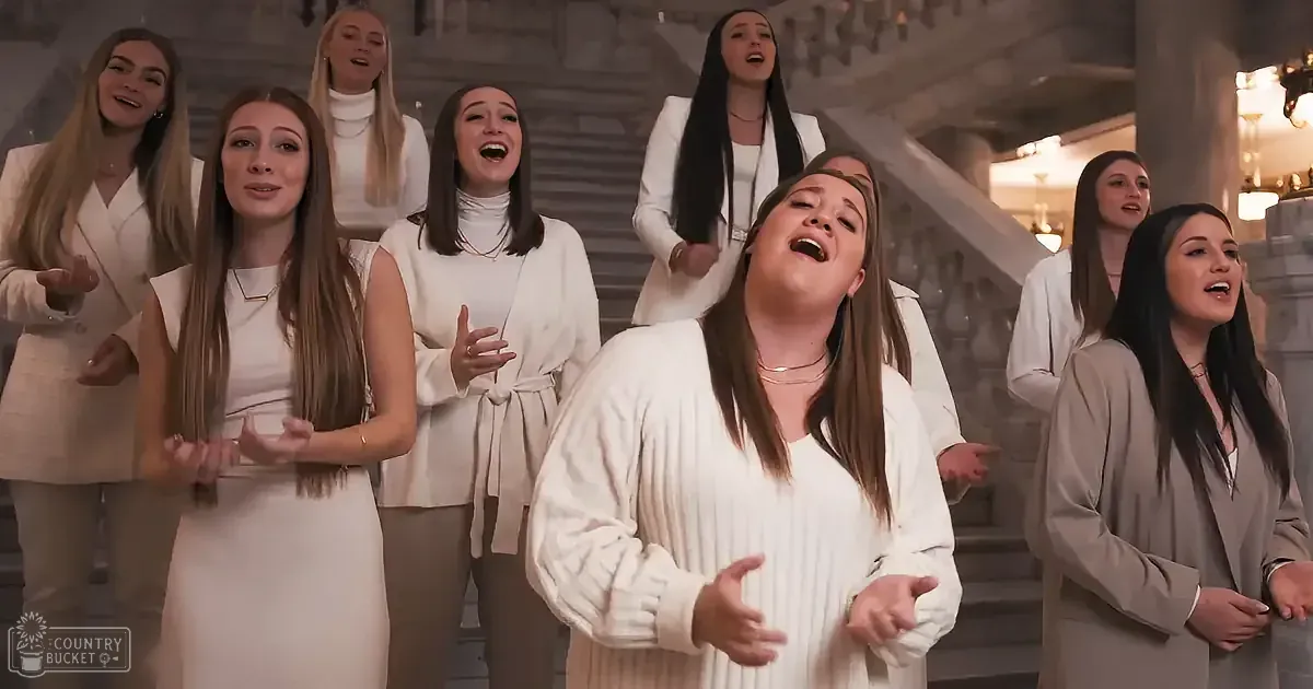 A woman in white leads a choir in song, surrounded by others in formal attire, in a grand, ornate setting.