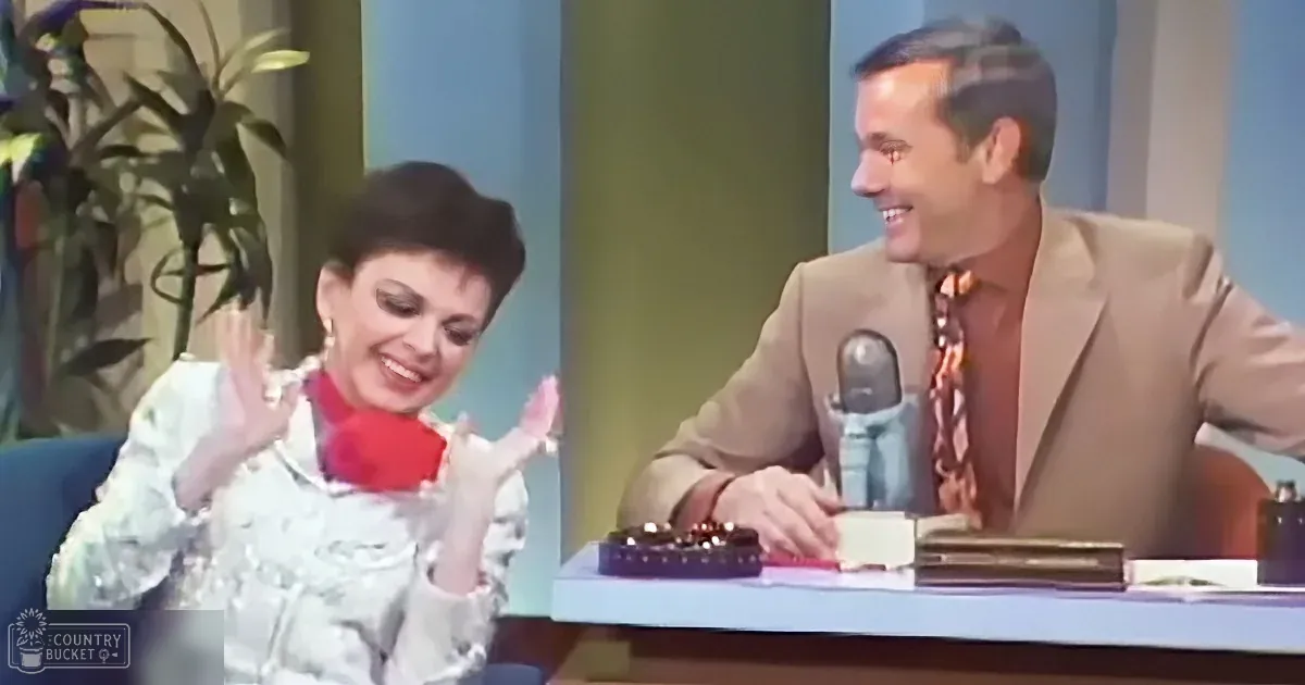 A woman in a white outfit and red scarf gestures animatedly while seated next to a man in a beige suit and patterned tie.