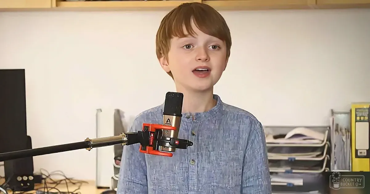 A young boy stands in front of a microphone, speaking in a room with shelves and equipment.