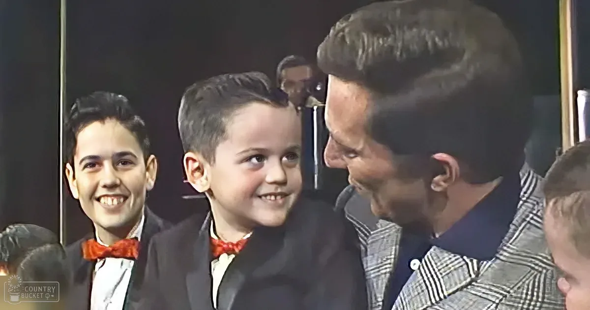 A young boy in a suit smiles at an older man in a checkered suit, surrounded by others in formal attire.