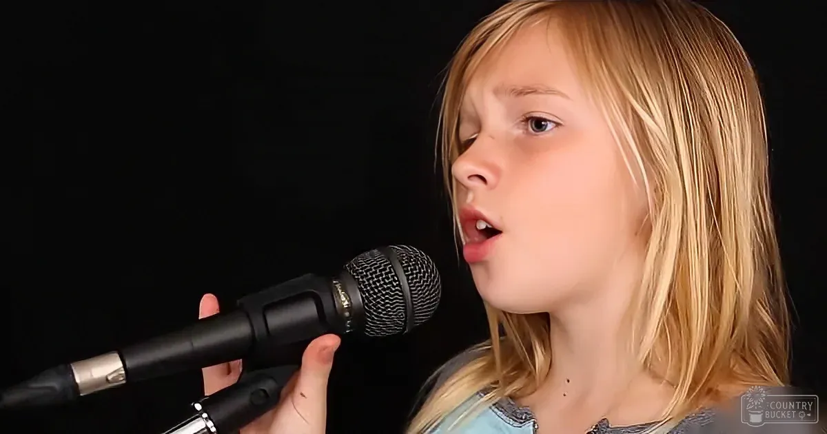 A young girl with blonde hair sings into a microphone against a dark background.