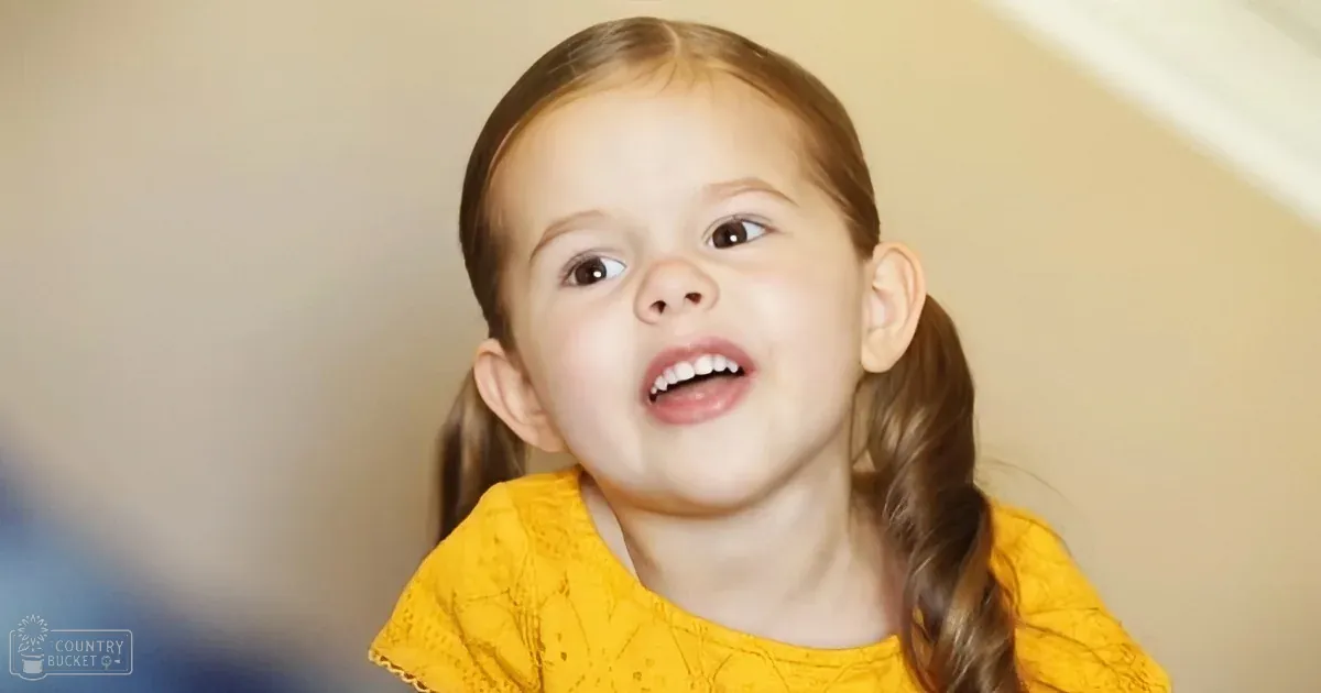 A toddler girl wearing a red knitted sweater stands indoors by a lit brick fireplace, with a gray upholstered chair and warm lighting, suggesting an evening winter setting.