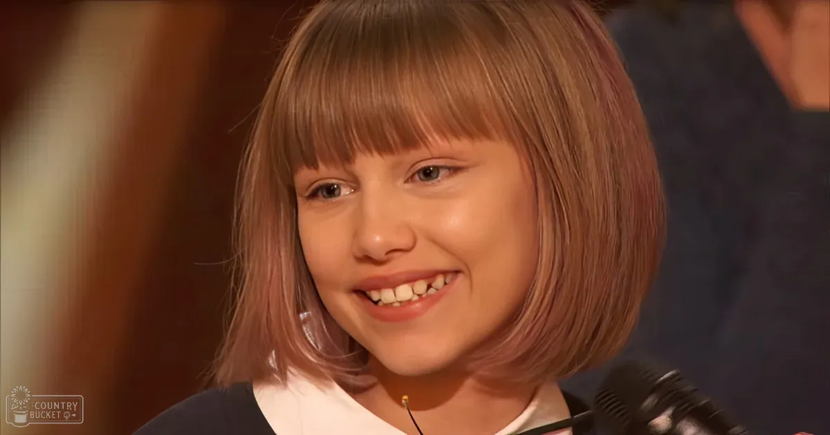 A young girl with short, light brown hair smiles while wearing a dark top, with a microphone visible in the foreground.