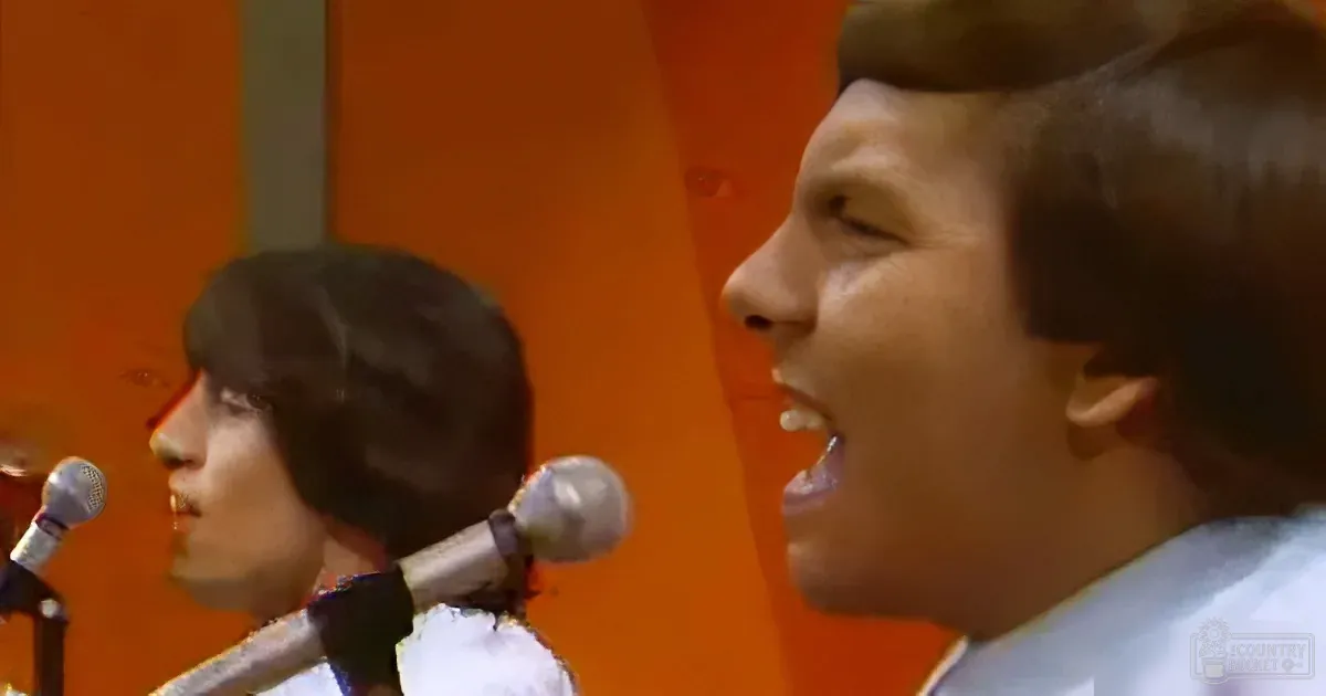 A young man with short hair sings into a microphone, with another person in the background.