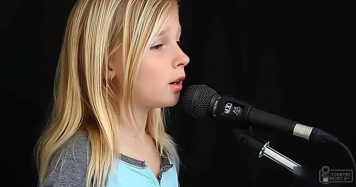 A young person with long blonde hair sings into a black microphone against a dark background.